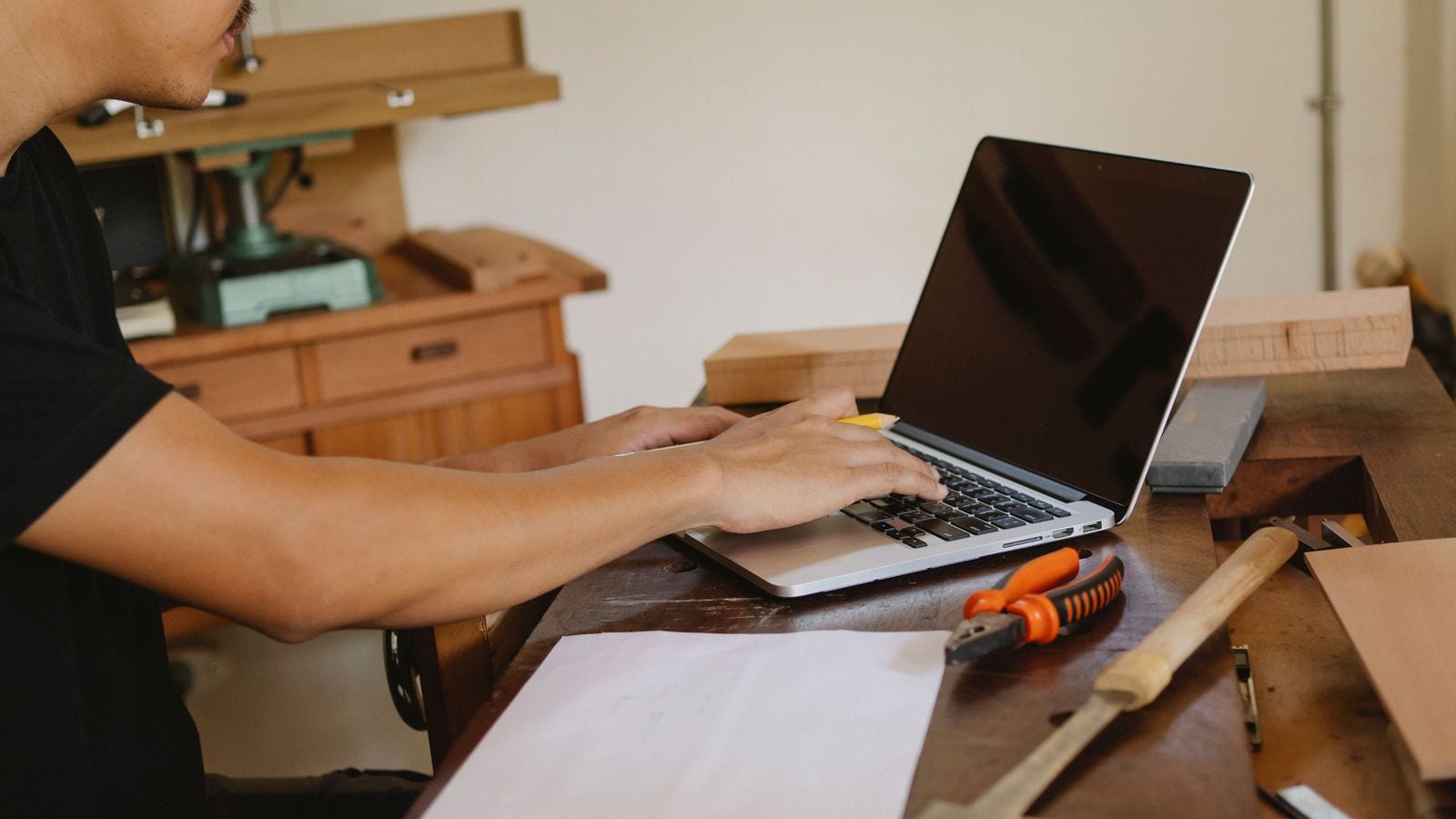 A tradesperson typing on a laptop at a workbench, with hand tools and offcuts beside it